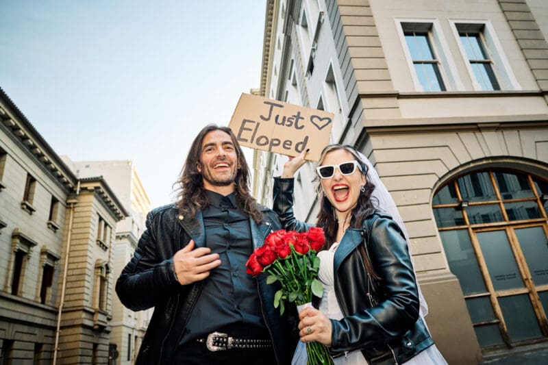 A cheerful couple showcases their spontaneous spirit in the city, the bride holding a bouquet of red roses, as they proudly display their 'Just Eloped' sign, their smiles as bright as the day.